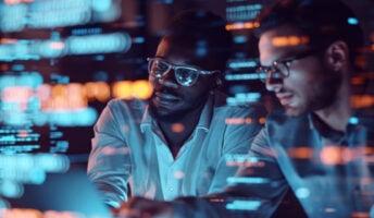 Two employees look at a big screen with data on it