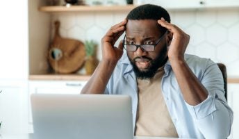A man with a horrified expression looks at a laptop, while sitting in the kitchen