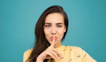 Woman in a yellow top on a turquoise background with her finger on her lips to signify quiet